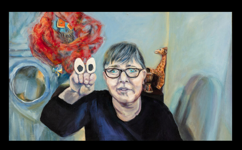 Woman with glasses holding her hand up with paper eyes on her knuckles as a finger puppet against a blue wall
